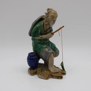 May include: A ceramic figurine of a fisherman wearing a green and brown robe, holding a fishing rod and sitting on a blue and white jar. The figurine is on a base with leaves.