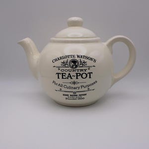 May include: Cream-colored ceramic teapot with a lid and handle. The teapot has black text that reads "CHARLOTTE WATSON'S COUNTRY TEA-POT For All Culinary Purposes". The teapot is from Henry Watson Pottery, England, founded in 1800.