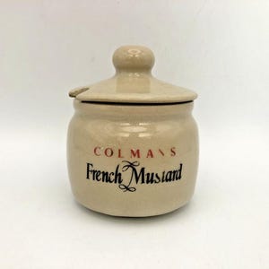 Może przedstawiać: Beżowy ceramiczny słoiczek na musztardę z pasującą pokrywką z małym wycięciem na łyżeczkę. Z przodu słoiczka znajduje się czerwony napis COLMAN'S nad czarnym napisem French Mustard wykonanym ozdobną czcionką, z gładkim, nakrapianym wykończeniem.