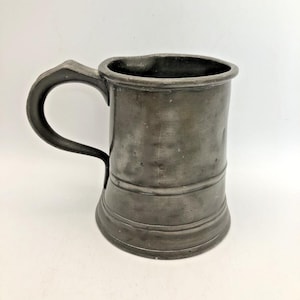 May include: A dark gray pewter mug with a smooth, matte finish and a curved handle. The cylindrical vessel features a slightly flared base and a wide rim, displaying a weathered, metallic texture with subtle surface variations and indentations.