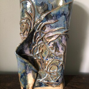 Può includere: Un vaso in ceramica alto fatto a mano con una silhouette irregolare e piegata. La superficie presenta uno smalto lucido nei toni del blu profondo, viola e bianco, accentuato da linee di argilla in rilievo che creano un motivo organico strutturato.