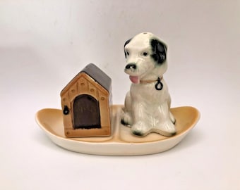 May include: A ceramic salt and pepper shaker set featuring a dog and dog house. The dog is white with black spots and a pink tongue. The dog house is brown with a black roof. The set sits on a cream-colored oval dish.
