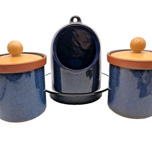 May include: A set of three ceramic kitchen containers. Two matching blue speckled jars with wooden lids and a small blue hanging container. The lids are light wood with a brown rim. The containers are on a black base.