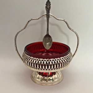 Könnte beinhalten: Eine tiefrote Glasschale in einem silberfarbenen Metallgestell mit filigranem Muster, einem Standfuß und einem hohen, geschwungenen Henkel. Ein kleiner silberfarbener Metalllöffel hängt oben am Henkel und ruht in der roten Glasschale.