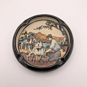 May include: A round ceramic ashtray with a black glazed rim and a central hand-painted illustration. The scene depicts a person kneeling to milk a goat in front of a house with a brown roof, set against a backdrop of rolling hills and a tree.