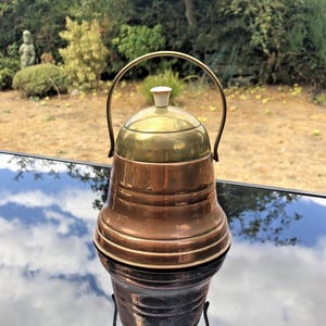 May include: A bell-shaped object constructed from copper with a brass dome top and a small white knob handle. A thin, arched brass handle is attached to the sides of the copper body, which features horizontal ridges around the lower section.