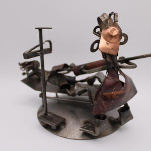 Puede incluir: Una escultura de metal hecha a mano que representa a un dentista y su paciente. La figura del dentista, con una cara de cobre, sostiene un clavo como herramienta. El paciente se reclina en una silla de metal. La escultura está montada sobre una base circular.