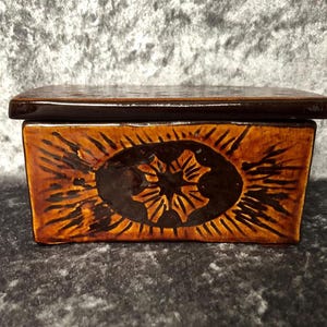 May include: A rectangular ceramic box with a dark brown lid and a body featuring a golden-brown glaze. The front panel displays a central dark brown oval containing a four-petaled floral motif surrounded by radiating dark brown etched lines.