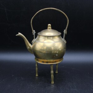 May include: A brass teapot with a handle and a spout, sitting on a three-legged stand. The teapot is a traditional design with a round body and a lid.