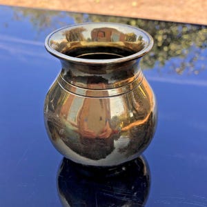 May include: A small, polished brass vessel with a rounded, bulbous body and a flared, circular rim. The metallic surface is highly reflective, showing a clear mirror-like finish that captures the surrounding environment in its golden sheen.