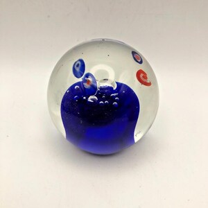 May include: A clear glass sphere paperweight featuring a deep blue base with suspended air bubbles. The upper portion of the sphere contains small, scattered circular accents in shades of blue and red, creating a distinct visual contrast within the transparent glass structure.