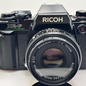 May include: A black Ricoh XR-P multi-program camera with a 50mm lens. The camera has a black body with silver accents and a prominent lens. The lens is marked "Made in Japan". The camera is designed for photography.