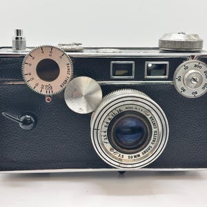 May include: A vintage Argus C3 rangefinder camera with a black textured body and silver metal accents. The lens is a 50mm f/3.5 Argus Coated Cintar. The front features a circular rangefinder dial, a shutter release lever, and a film advance knob.