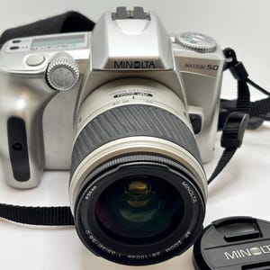 Könnte beinhalten: Eine silberne Minolta Maxxum 50 35mm Filmkamera mit schwarzem Trageriemen und abnehmbarem Objektivdeckel. Das Kameragehäuse hat ein silbernes Finish, einen schwarzen gummierten Zoomring am Objektiv und den Schriftzug Minolta Maxxum 50 vorne.