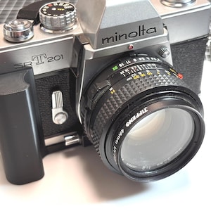 Può includere: Una fotocamera a pellicola Minolta SR T 201 da 35 mm argentata e nera con un obiettivo nero. L'obiettivo ha un filtro UV Tiffen da 49 mm. La fotocamera ha un'impugnatura nera e vari quadranti e pulsanti.