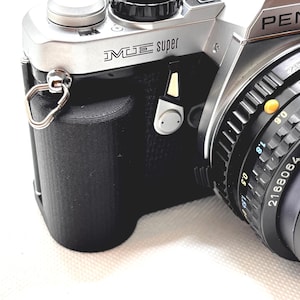 May include: Close-up of a silver and black Pentax ME Super 35mm film camera. The camera body is black with silver accents, and the lens is black with measurement markings. The camera has a strap attachment and the word "PENTAX" is visible.