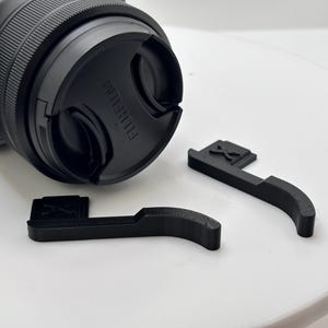 Könnte beinhalten: Schwarze Objektivkappe mit Fujifilm-Logo und zwei schwarzen Kamera-Daumengriffen. Die Daumengriffe haben ein X-Design. Das Kameraobjektiv ist schwarz und hat ein geriffeltes Design.