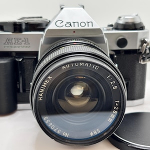 Puede incluir: Una cámara Canon AE-1 Program con una lente Hanimex Automatic 1:2.8 f=28mm. El cuerpo de la cámara es plateado y negro, con el logotipo de Canon en la parte superior. La lente tiene un diámetro de filtro de 58 mm.
