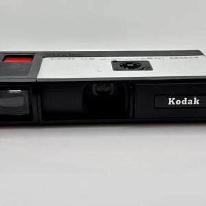 Könnte beinhalten: Eine schwarz-silberne Kodak Pocket Instamatic 20 Kamera mit rechteckigem Gehäuse, einem Sucher auf der linken Seite, einem zentralen Objektiv und einem schwarzen Auslöser mit rotem Akzent. Der Markenname Kodak ist weiß auf der Vorderseite aufgedruckt.