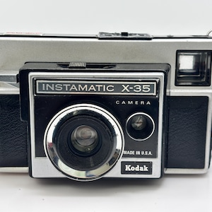 May include: A vintage Kodak Instamatic X-35 camera. The camera has a silver top and black front with the text "Instamatic X-35 Camera" and "Made in U.S.A." The lens is surrounded by a silver ring. A black strap is attached.