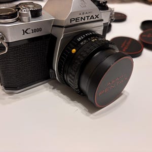 Può includere: Una fotocamera Asahi Pentax K1000 argento e nera con un obiettivo nero e un copriobiettivo. Il copriobiettivo riporta le parole "Asahi Pentax" in rosso. Diversi copriobiettivo neri con finiture rosse sono sullo sfondo.