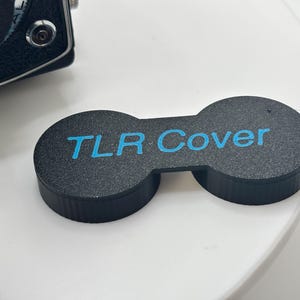 Könnte beinhalten: Ein schwarzer, texturierter, doppelt kreisförmiger Objektivdeckel für eine zweiäugige Spiegelreflexkamera. Das Objekt trägt den Text "TLR Cover", der in leuchtendem Cyanblau auf dem Mittelsteg zwischen den beiden Kappen gedruckt ist.