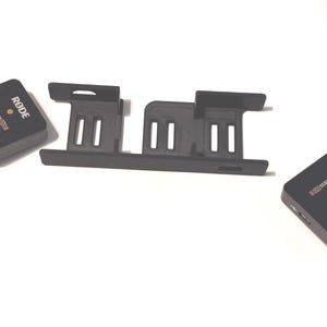 Może przedstawiać: Czarne urządzenia audio, w tym dwa małe prostokątne urządzenia z logo "RØDE WIRELESS GO II" i czarny plastikowy wspornik z otworami, są wyświetlane na białej powierzchni.