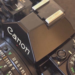 May include: Close-up of a black Canon camera. The camera has a lens with white numbers and markings, and the word "Canon" is printed in white on the top. The camera also has a flash attachment.