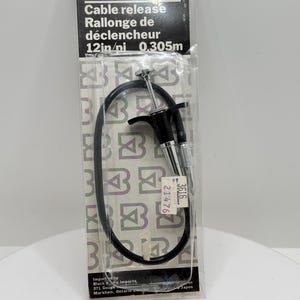 Puede incluir: Un disparador de cable negro para cámaras en su empaque original de plástico transparente. La etiqueta dice Black's Cable release, 12in/ni 0.305m. El dispositivo cuenta con un cable negro flexible con un mecanismo de émbolo de metal plateado.