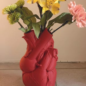 Könnte beinhalten: Eine rote, herzförmige Vase mit einem Blumenstrauß. Die Blumen sind gelbe Lilien, grüne Chrysanthemen und eine rosa Nelke. Die Vase hat ein detailliertes anatomisches Design.