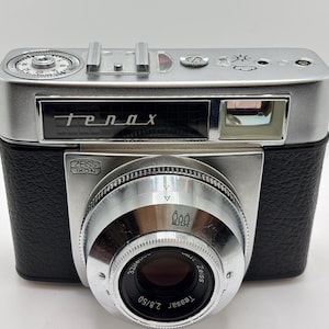 May include: A silver and black Zeiss Ikon Tenax camera featuring a prominent lens barrel with a Carl Zeiss Tessar 2.8/50 lens. The top plate displays a film advance dial, a hot shoe, and weather icons. The front includes the Tenax logo and branding.