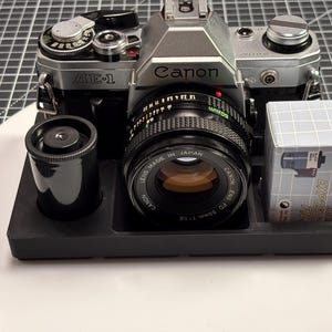 Könnte beinhalten: Eine silberne und schwarze Canon AE-1 Filmkamera mit einem 50-mm-Objektiv, einer Filmkartusche und einer Filmbox. Die Kamera befindet sich auf einem schwarzen Tablett. Auf dem Objektiv steht "MADE IN JAPAN".