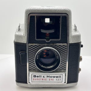 May include: A vintage Bell & Howell Electric Eye 127 camera. The camera has a silver top and a black body with a textured pattern. The lens is black with red and white details. The camera has the words "Bell & Howell" and "Electric Eye 127" on the front.