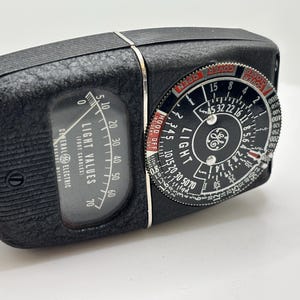 May include: A vintage General Electric light meter, black with a textured finish. The meter features a dial with numbers and markings, including "LIGHT VALUES" and "GENERAL ELECTRIC". A smaller dial indicates light settings.