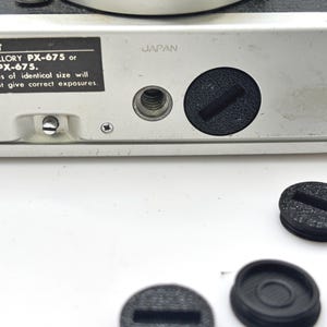 Puede incluir: Primer plano de la base plateada de una cámara con la palabra "JAPAN" impresa. La base tiene un compartimento para la batería y una tapa negra. También se ve el texto "IMPORTANTE" e información sobre la batería.