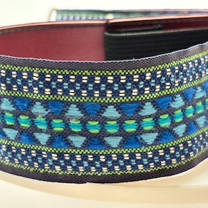 May include: Close-up of a woven strap with a repeating pattern of blue, green, and white geometric shapes. The strap has a dark blue border and a burgundy interior. The design is intricate and detailed, showcasing a blend of colors and textures.