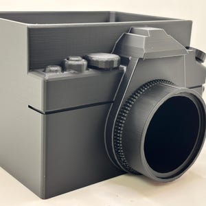 May include: A black, 3D-printed desk organizer shaped like a vintage camera. The organizer features a lens, buttons, and a viewfinder, with a rectangular compartment for storage. The design is a solid, matte black.