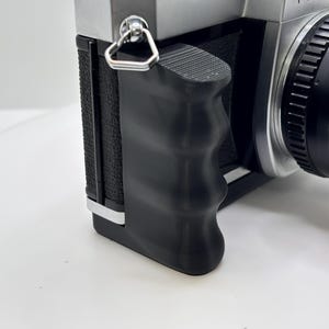 Puede incluir: Primer plano de una cámara vintage plateada y negra. La cámara tiene una empuñadura negra, una lente y un cuerpo plateado. La empuñadura tiene textura para un agarre seguro. La lente tiene múltiples anillos para el enfoque y la apertura.