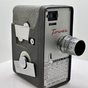 Puede incluir: Una cámara de película Tower Model T.92 de 8 mm vintage. La cámara es gris con una lente plateada y una tapa de lente negra. La palabra "Tower" está en escritura roja. La cámara tiene una tabla con ajustes de exposición.