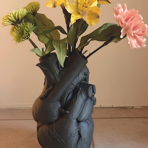 Könnte beinhalten: Eine schwarze, herzförmige Vase mit einem Blumenstrauß. Die Blumen sind gelbe Lilien, grüne Chrysanthemen und eine rosa Nelke. Die Vase weist anatomische Details auf.