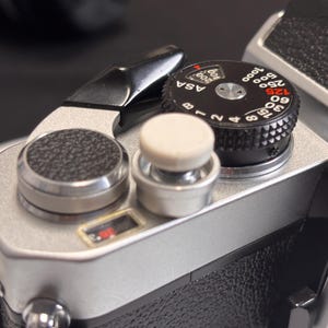 May include: Close-up of a vintage camera's top plate, showcasing dials and controls. The silver body contrasts with black dials and a textured grip. A white button and a red indicator are also visible. The ASA dial is marked with numbers.