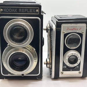 May include: A black Kodak Reflex camera with dual lenses and silver dials sits next to a smaller black Kodak Duaflex II camera. Both vintage cameras feature metallic accents, visible lens markings, and text labels on their front panels.