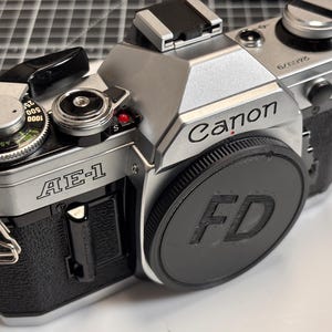 May include: Silver and black Canon AE-1 film camera with a black lens cap marked "FD." The camera has various dials and settings visible, including an ASA dial and a shutter speed dial. The Canon logo is prominently displayed.