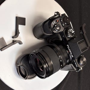 Puede incluir: Cámara Fujifilm X-S20 negra con lente y tapa de lente, sobre una superficie circular blanca. La lente está marcada 16-50mm. Accesorios negros adicionales se colocan cerca de la cámara.