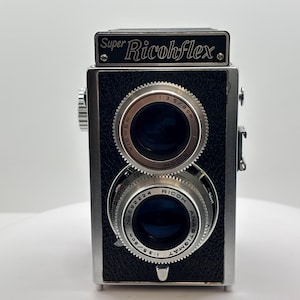 Peut inclure: Un appareil photo reflex bi-objectif Super Ricohflex vintage avec un corps noir texturé et des accents en métal argenté. L'avant présente deux objectifs circulaires à bords argentés, marqués Ricoh Anastigmat 1:3.5/8cm et Viewer 1:3.5/8cm.