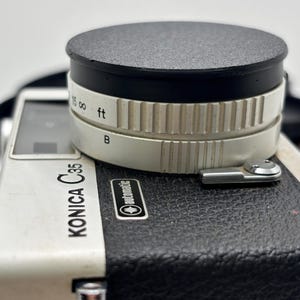 Könnte beinhalten: Eine Vintage Konica C35 Kamera mit einem schwarzen und silbernen Gehäuse. Die Kamera hat einen schwarzen Objektivdeckel und einen silbernen Objektivring mit Entfernungsmarkierungen in Fuß. Die Kamera befindet sich auf weißem Hintergrund.