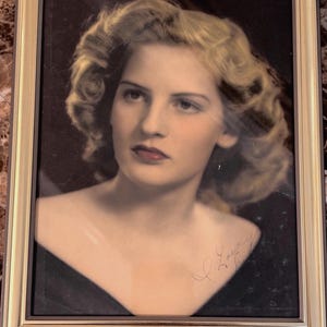 May include: A framed portrait of a woman with blonde hair, wearing a black dress. The portrait is in a gold-colored frame. The woman has red lipstick and a signature in the bottom right corner.