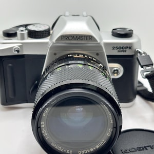 以下が含まれることがあります： シルバーとブラックのPromaster 2500PK Super 35mmフィルムカメラ。取り外し可能なレンズには35-70mm 1:3.5-4.5 C/D Zoom+Macroと刻印されています。カメラ本体には黒いグリップ、上部のダイヤル、側面に黒いストラップが付いています。
