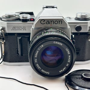 Puede incluir: Una cámara Canon AE-1 plateada y negra con una lente de 50 mm. La lente está etiquetada como "CANON LENS FD 50mm 1:1.8 MADE IN JAPAN". La cámara tiene una correa negra y una tapa de lente.