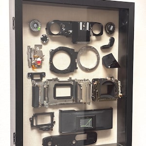 May include: A framed display of dismantled camera parts, including lenses, body components, and internal mechanisms. The black frame encloses the components against a neutral background, showcasing the intricate details of the camera's internal structure.